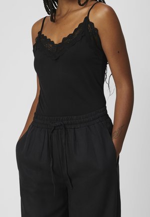 Kvinde iført sort camisole med blondekant og løse sorte bukser med snøre i taljen, hænderne i lommerne, der viser tatovering på armen og flettet hår.