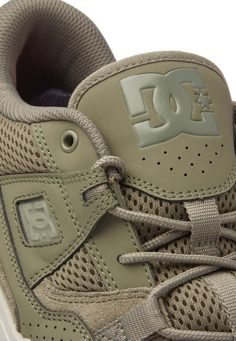 Zapatilla verde con parte superior de malla y sintético suave, que presenta un logo prominente en la lengüeta y detalles perforados para ventilación.