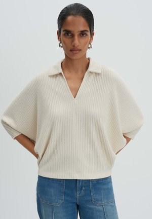 Vrouw met donker haar, met zilverkleurige oorringen, een crèmekleurige geribbelde top met kraag en een blauwe spijkerbroek, staand met handen in de zij.