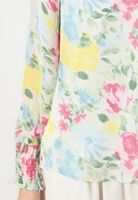 Blouse fleurie en tissu léger et transparent avec des roses pastel et des feuilles en rose, jaune et bleu. Détails de poignets froncés sur les manches.