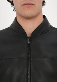 Jachetă din piele neagră, cu guler în picioare, închidere cu fermoar și un finisaj mat, neted. Detalii cusute orizontal pe piept.