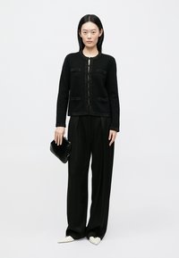 Černá pletená bunda s texturovanými doplňky, kulatým výstřihem a předními kapsami, kombinovaná s širokými černými kalhotami a černou kabelkou.