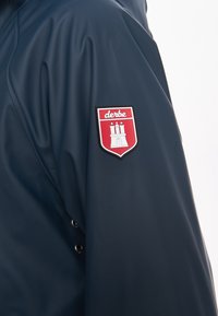 Mouw van een marineblauw jack met een rode en witte Derbe-merkembleem waarop een kasteel met drie torens en sterren erboven staat.