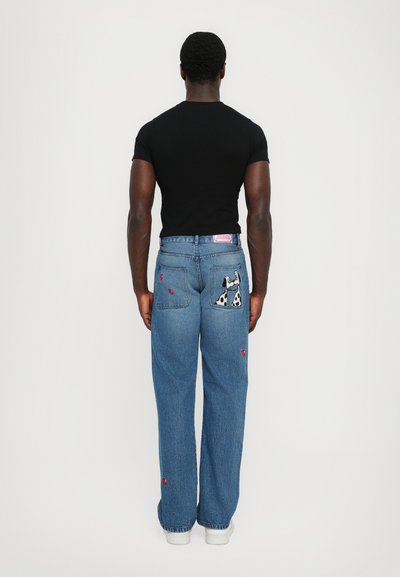 Jeans de denim albastru largi, cu patch-uri în formă de inimă roșie și un grafic cu Dalmațian pe buzunarul din spate. Purtați cu un tricou negru bine ajustar.