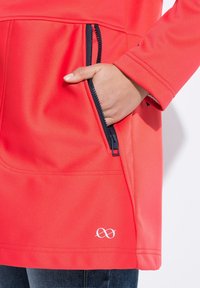 Rode softshell jas met een ritszak, contrasterende marineblauwe rits en een decoratieve logo nabij de zoom. Close-up van de zakdetails.