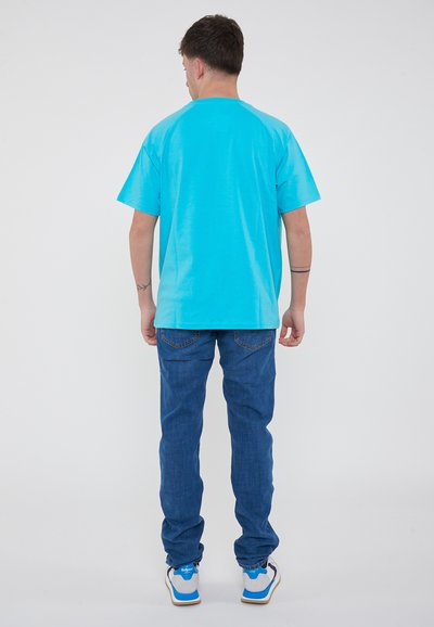 T-shirt in cotone turchese con maniche corte, abbinato a jeans in denim blu. Sneakers bianche e blu completano l'outfit.