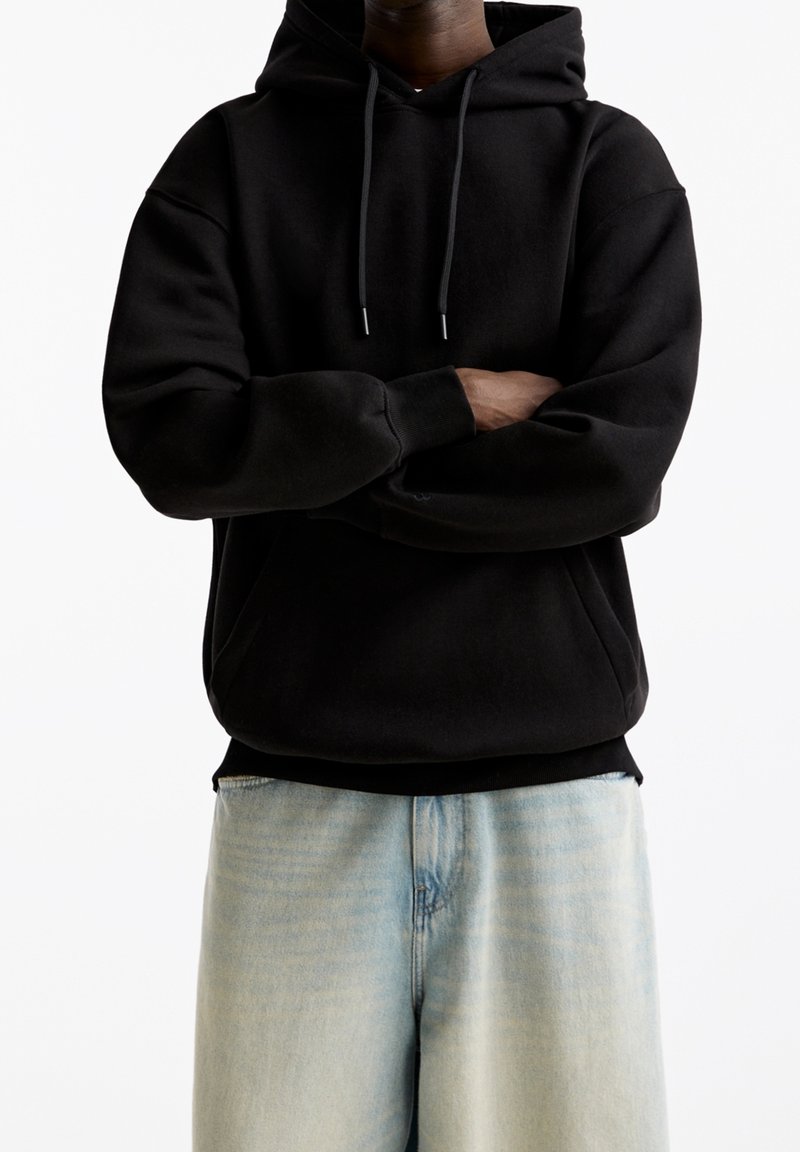 Personne portant un sweat à capuche noir avec des cordons et un jean bleu clair, les bras croisés sur la poitrine, sur un fond blanc uni.