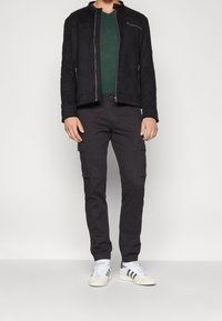 Giacca di suede nera, maglietta verde scuro a V, pantaloni cargo neri con tasche laterali e sneaker bianche con strisce nere. Design semplice e moderno.