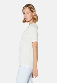 Weißes, kurzärmliges T-Shirt aus weichem Stoff, mit rundem Ausschnitt und Spitzeneinsatz sowie seitlichen Schlitzen. Getragen mit hell gemusterten Hosen.