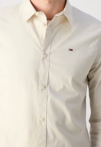 Camicia button-up beige chiaro realizzata in tessuto liscio, con colletto classico e un piccolo logo accentato sul lato sinistro del petto.