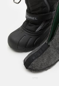 Juodas izoliuotas botas su guminėmis padais ir tekstiliniu viršumi, derinamas su pilku vilnonio pamušalu, kuriame yra juodas audinio juostas, užtikrinanti tvirtą prigludimą.