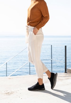 Zapatos deportivos negros con tela texturizada, combinados con pantalones beige claro con cinturón de atar y un suéter de punto marrón con costuras intrincadas.