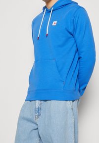 Sudadera azul de tejido suave, con un bolsillo canguro, cordones blancos con puntas rojas y un pequeño parche de logo en el pecho.