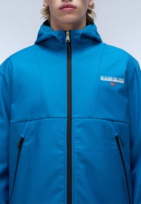 Blå udendørsjakke med zip-lukning, hætte og sidelommer. Har en logopatch og kontrasterende sort lynlås foran.