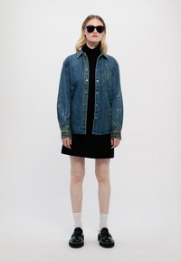 Jachetă din denim de culoare albastră decolorată, cu stropi de vopsea, purtată peste un pulover negru cu guler înalt și o rochie neagră, asortată cu pantofi negri și șosete albe.