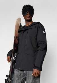 Άνδρας σε μαύρη σκι jacket και γκρι παντελόνι κρατά σκι, φορά μεγάλα μαύρα γυαλιά σκι σε απλό φόντο.