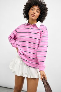 Polo a maniche lunghe a righe rosa con colletto bianco, caratterizzato da strisce orizzontali nere e un piccolo logo rosso. Abbinato a una gonna bianca.