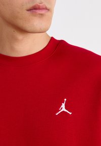 Červená bavlnená mikina s okrúhlym výstrihom a ribbenými manžetami, s bielym logom Jumpman na ľavej hrudi. Hladká textúra.