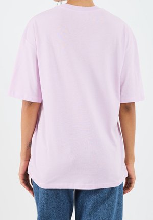 Person trägt ein hellviolettes, übergroßes T-Shirt mit kurzen Ärmeln und blaue Jeans, von hinten vor weißem Hintergrund zu sehen.