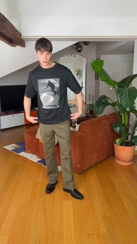 Mladi moški v črni majici z grafičnim potiskom in olivno zelenih hlačah stoji v dnevni sobi z lesenim tlemi, rjavo zofa, veliko rastlino in televizijo.