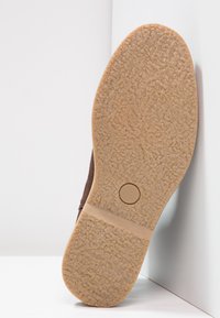 Braune Wildlederschuhe mit einer Gummisohle, die eine strukturierte, rutschfeste Oberfläche und ein kreisförmiges Akzentfeld im Fersenbereich aufweist.