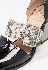 Chaussure en cuir verni noir avec une large sangle en imitation serpent texturée et une boucle en argent, mettant en avant un design moderne et des matériaux uniques.