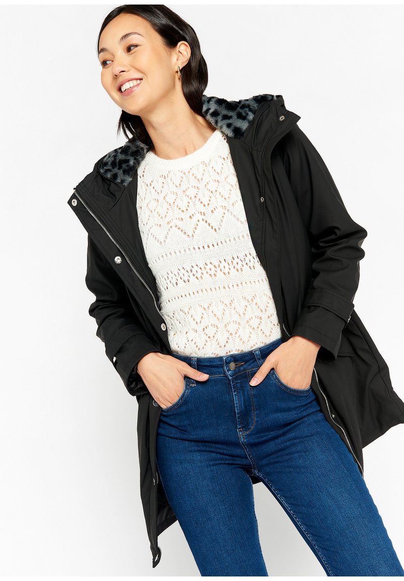 Zwarte waterdichte jas met een leopardprint nepbont kraag, gecombineerd met een witte gebreide trui en blauwe high-waisted jeans.