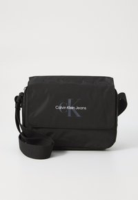 Borsa a tracolla nera in nylon con chiusura a patta, dotata del logo "Calvin Klein Jeans" in bianco e una tracolla regolabile con texture.