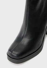 Schwarze Leder-Stiefelette mit glatter Textur, abgerundeter Zehenpartie, leichtem Plateau und tiefem, klobigem Absatz. Minimale Nähte setzen Akzente.