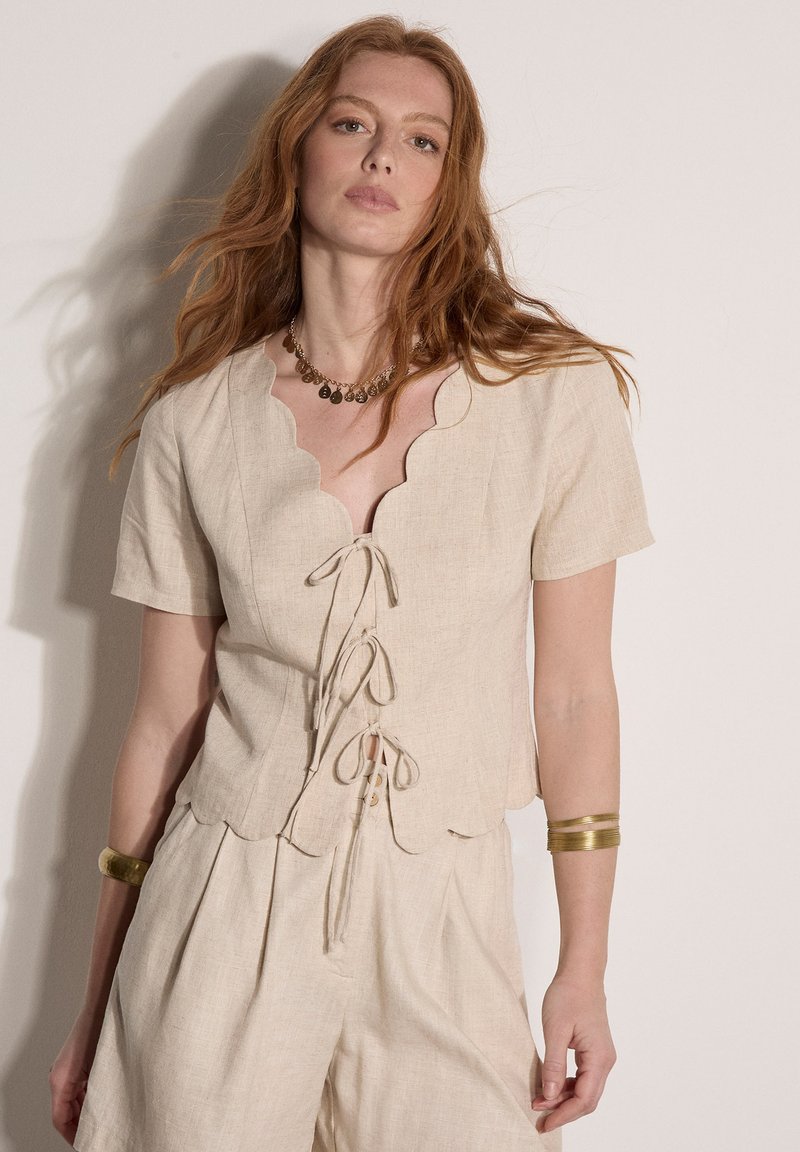 Femme aux cheveux roux portant un haut beige à manches courtes avec bords festonnés, noué devant, pantalon taille haute assorti, collier en or et bracelets.
