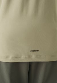 Camiseta deportiva de color verde oliva claro con textura suave, que presenta un logo negro "AEROREADY" en el dobladillo y detalles de costura limpios.