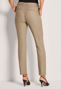 Vrouw in beige geruite slim-fit broek en zwarte hoge slingback schoenen, staand met handen in haar zakken.