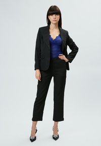 Zwarte op maat gemaakte blazer over een blauwe kanten body, gecombineerd met zwarte op maat gemaakte broeken. Zilverkleurige zwarte hakken maken de outfit compleet.