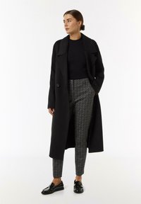 Black long coat with a collar, over a fitted black sweater and patterned trousers. The pants feature a geometric design, paired with glossy black loafers.