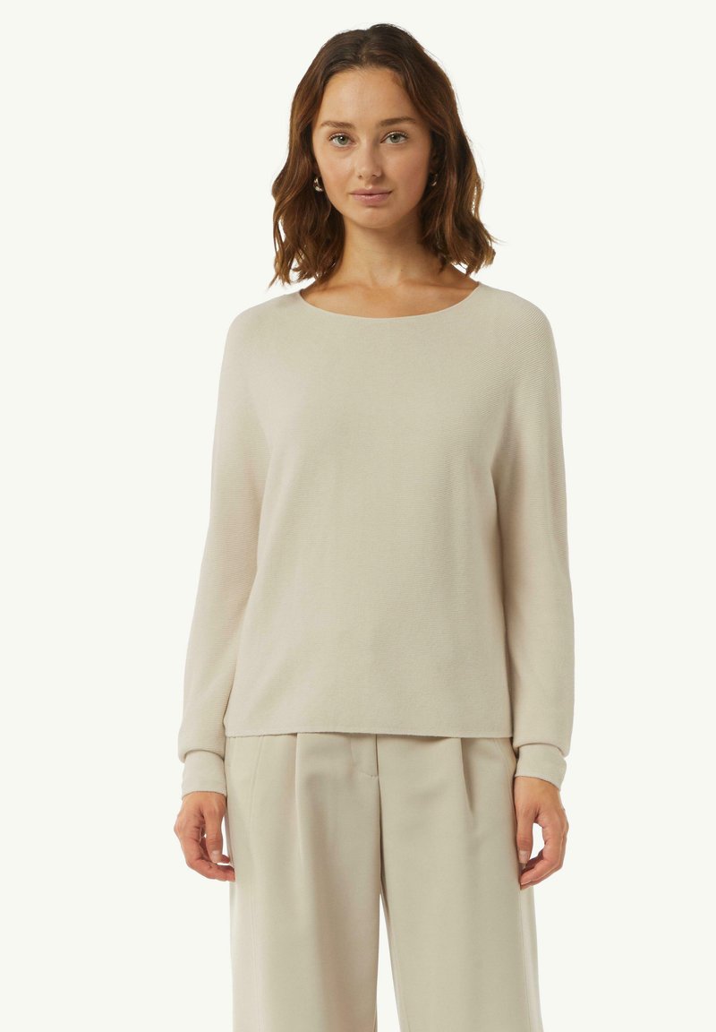 Helles beige langärmeliges Strickpullover mit weitem Rundhalsausschnitt und gerippter Textur. Er hat einen legeren Schnitt und ist mit passenden Hosen kombiniert.