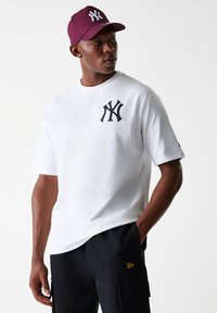 Camiseta blanca con logo negro de NY en el pecho, combinada con pantalones cortos cargo negros. Modelo lleva una gorra burdeos con emblema blanco de NY.