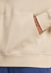 Beige waffle-textured hoodie featuring a front pocket and ribbed cuffs. The fabric is soft and slightly stretchy, with visible stitching details.