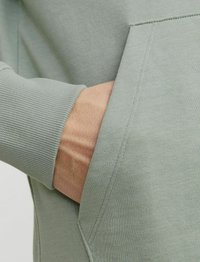Tessuto di felpa verde chiaro a coste con tasca frontale. Primo piano di una mano appoggiata nella tasca, che mette in evidenza la texture e i dettagli delle cuciture.