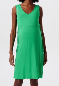 Vestido de maternidad verde en tejido suave, sin mangas, con escote en V, lazo ajustable en la cintura y un corte holgado.
