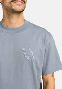 Man in een lichtblauw T-shirt met ronde hals en een geborduurd "VA"-logo op de linkerborst, zichtbare arm-tattoo.