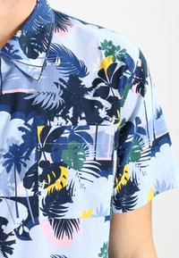 Kurzarmhemd mit blauem Hintergrund und lebhaften tropischen Pflanzenmustern, darunter dunkle Blätter und gelbe Akzente, mit einer Brusttasche.