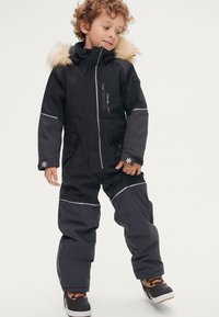 Zwarte geïsoleerde sneeuwoverall met een met bont gevoerde capuchon, reflecterende accenten, ritszakken en elastische manchetten. Gecombineerd met zwart en bruine laarzen.