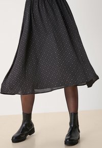 Žena v černých kotníkových botách, průhledných černých punčochách a volné černé sukni s malými bílými puntíky, stojící na světlé podlaze.