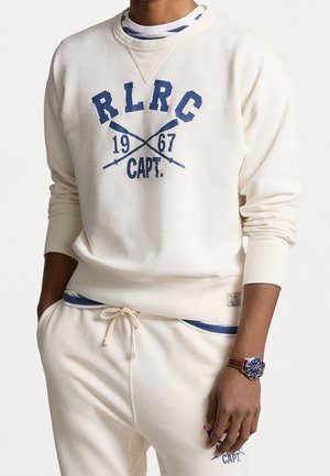 Felpa crema con testo blu "R.L.R.C. 1967 CAPT." e remi incrociati; polsini a coste; abbinata a pantaloni jogger coordinati. Orologio al polso.
