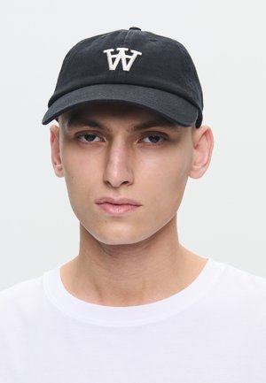 Ung mann iført en svart baseballcap med hvit "W"-logo og en enkel hvit t-skjorte med rund hals mot en lys bakgrunn.