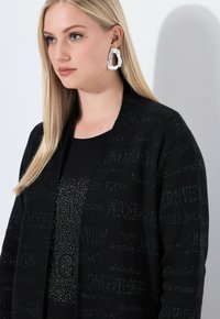 Černý cardigan s texturovaným vzorem písmen. Vnitřní košile má třpytivý design. Modelka má dlouhé vlasy a ozdobný náušník.