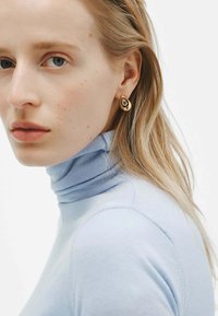 Pendiente en forma de lágrima dorado con una piedra central clara. Top de cuello alto azul claro, de textura suave, con un diseño ajustado. Estilo minimalista.