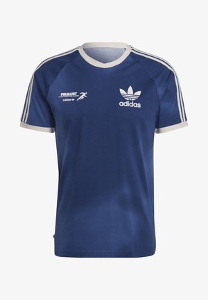 T-shirt Adidas bleu avec bordure et logo blancs, associé à un pantalon beige. Le mannequin porte des baskets blanches avec des rayures bleu marine et des accents beige.