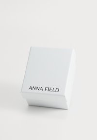Bijela kvadratna kutija s glatkom površinom, na kojoj se nalazi tekst "ANNA FIELD" u crnoj boji. Jednostavan dizajn bez dodatnih akcenata ili uzoraka.