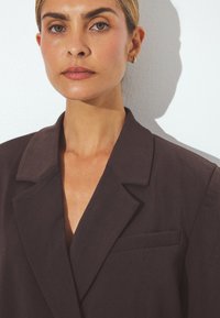 Ciemnobrązowa marynarka z klapami w kształcie notcha, jedną kieszenią i zorganizowaną konstrukcją. Gładki materiał o subtelnym połysku i prostych liniach.